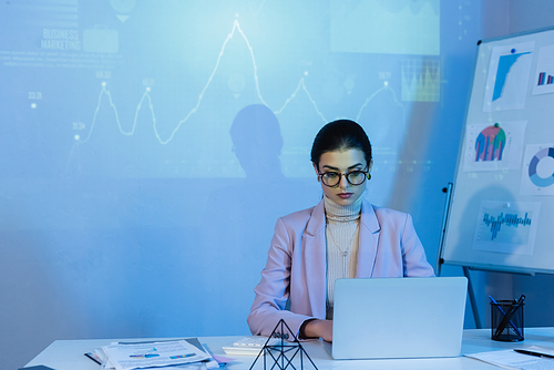businesswoman in glasses using laptop near digital graphs on wall