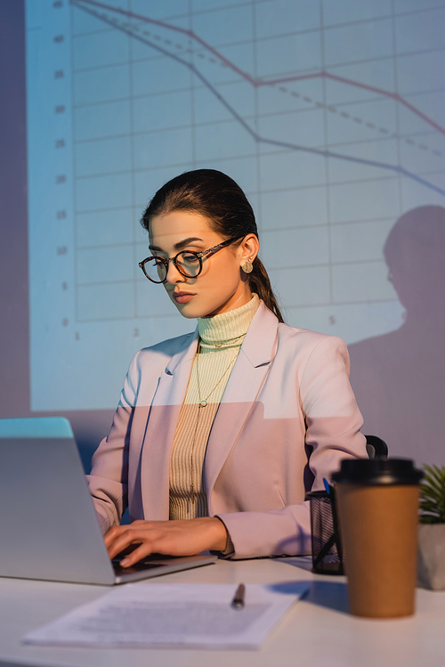 businesswoman in glasses using laptop near digital graphs on wall and paper cup on blurred foreground