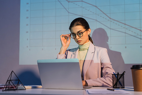 businesswoman adjusting glasses and using laptop near digital graphs on wall and paper cup on blurred foreground