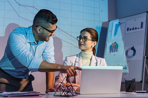 happy businessman pointing with finger at laptop and looking at coworker in glasses