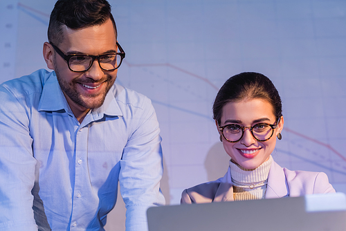 cheerful business people in glasses looking at laptop near digital charts on wall