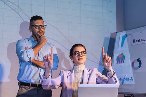 businesswoman in glasses pointing with fingers near businessman in glasses looking at digital graphs