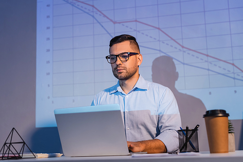 businessman in glasses looking at laptop near graphs on wall and paper cup on desk