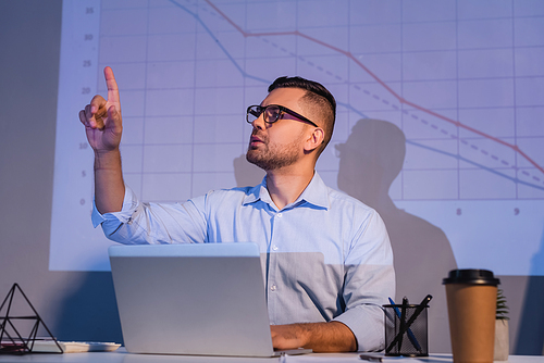 businessman in glasses pointing with finger near laptop and graphs on wall