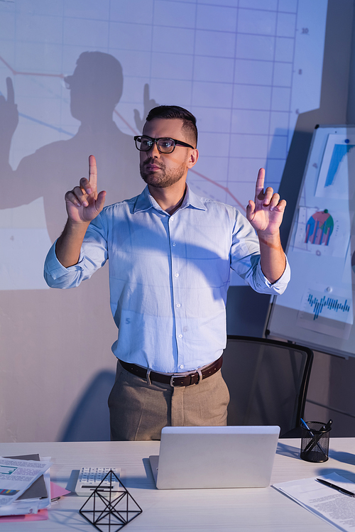 businessman in glasses while pointing with fingers near digital graphs in office
