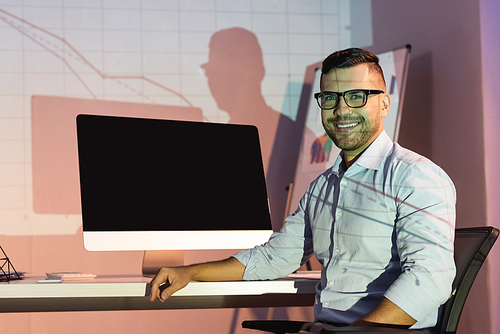 happy businessman in glasses  while sitting near computer monitor with blank screen