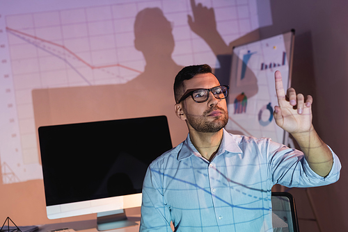 businessman in glasses pointing with finger near computer monitor with blank screen in office