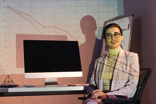 happy businesswoman in glasses  while sitting near computer monitor with blank screen