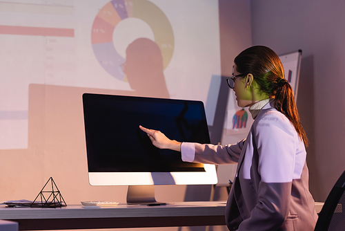 businesswoman in glasses pointing with finger at computer monitor with blank screen in office