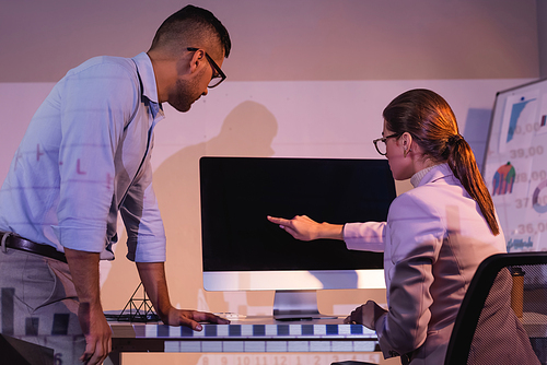 businesswoman in glasses pointing with finger at computer monitor with blank screen near coworker in office