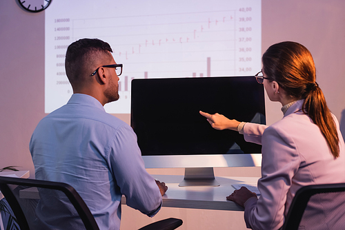 businesswoman in glasses pointing with finger at computer monitor with blank screen near coworker in office