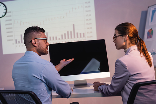 businessman in glasses pointing with hand at computer monitor with blank screen near coworker in office