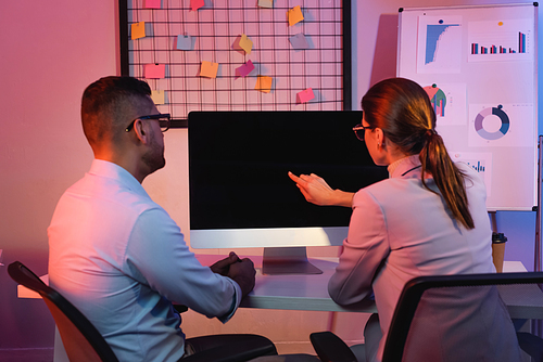 businesswoman pointing with finger at computer monitor with blank screen near coworker in office