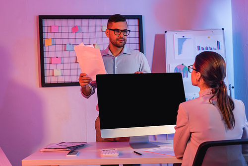 businesswoman sitting near computer monitor with blank screen and looking at coworker with documents