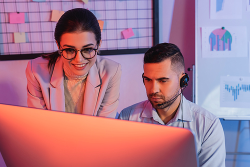 operator in headset looking at computer monitor near smiling businesswoman