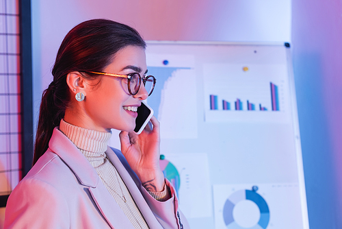 cheerful businesswoman talking on smartphone near flipchart with charts and graphs