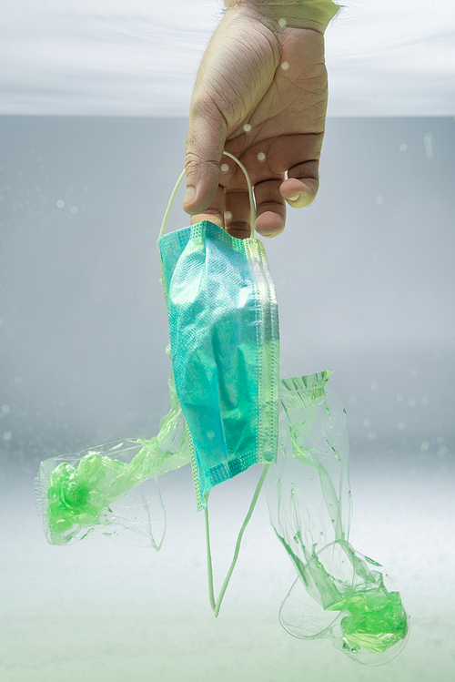 partial view of male hand near medical mask and plastic rubbish in water, ecology concept