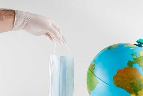 cropped view of man in latex glove holding medical mask near globe isolated on grey, ecology concept