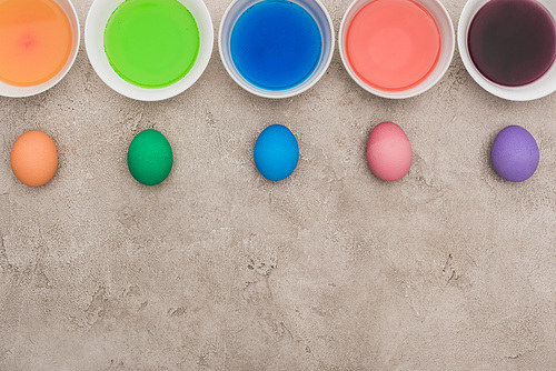 top view of painted chicken eggs near watercolor paints in bowls on grey concrete surface