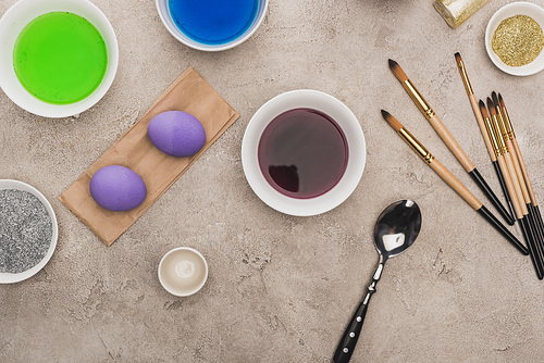 top view of chicken eggs, glitter and watercolor paints with paintbrushes on grey concrete surface