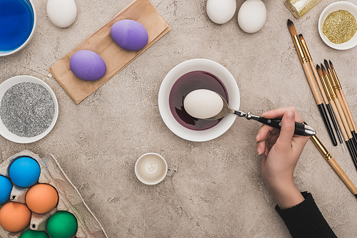 cropped view of woman coloring chicken eggs in watercolor paints on grey concrete surface