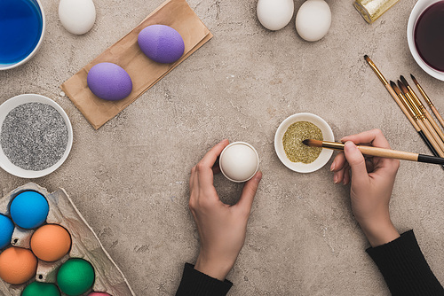 cropped view of woman decorating chicken eggs with golden glitter on grey concrete surface