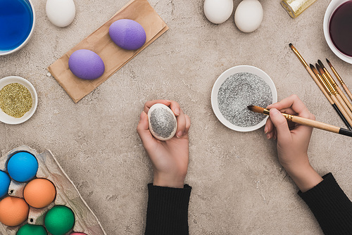 cropped view of woman decorating chicken eggs with silver glitter on grey concrete surface