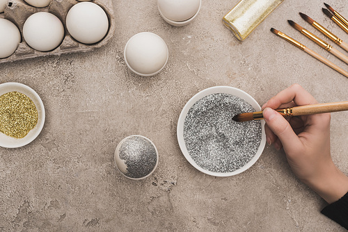 cropped view of woman decorating chicken egg with silver glitter on grey concrete surface