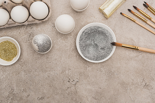 top view of chicken eggs, silver and golden glitter with paintbrushes on grey concrete surface