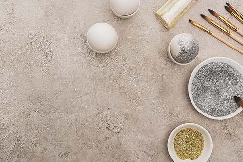 top view of chicken eggs, silver and golden glitter with paintbrushes on grey concrete surface