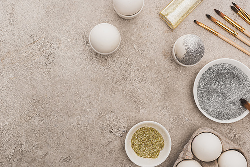 top view of chicken eggs, silver and golden glitter with paintbrushes on grey concrete surface