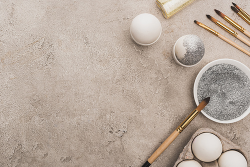 top view of chicken eggs, silver and golden glitter with paintbrushes on grey concrete surface