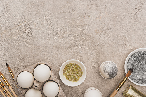 top view of chicken eggs, silver and golden glitter with paintbrushes on grey concrete surface