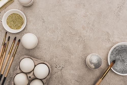 top view of chicken eggs, silver and golden glitter with paintbrushes on grey concrete surface