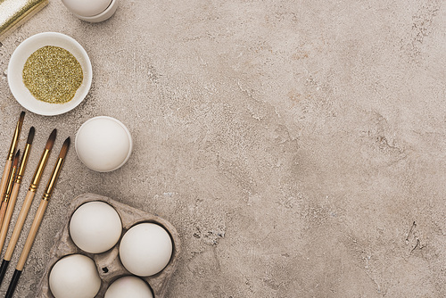 top view of chicken eggs, golden glitter with paintbrushes on grey concrete surface