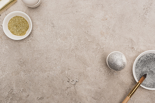 top view of chicken eggs, silver and golden glitter with paintbrushes on grey concrete surface