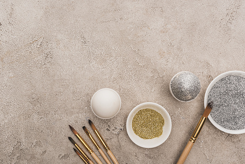 top view of chicken eggs, silver and golden glitter with paintbrushes on grey concrete surface