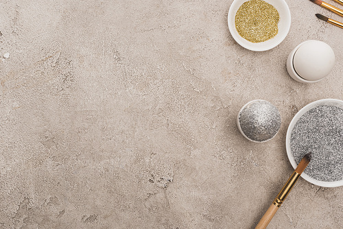 top view of chicken eggs, silver and golden glitter with paintbrushes on grey concrete surface