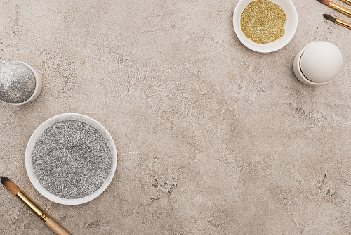 top view of chicken eggs, silver and golden glitter with paintbrushes on grey concrete surface