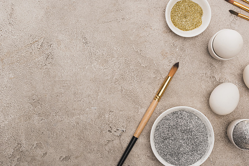 top view of chicken eggs, silver and golden glitter with paintbrushes on grey concrete surface