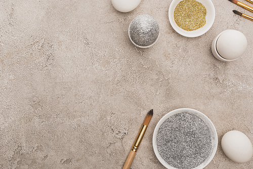 top view of chicken eggs, silver and golden glitter with paintbrushes on grey concrete surface