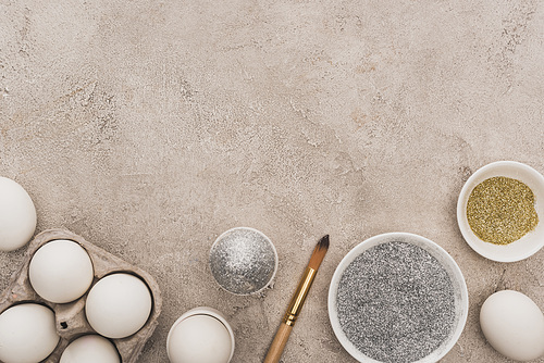 top view of chicken eggs, silver and golden glitter with paintbrushes on grey concrete surface