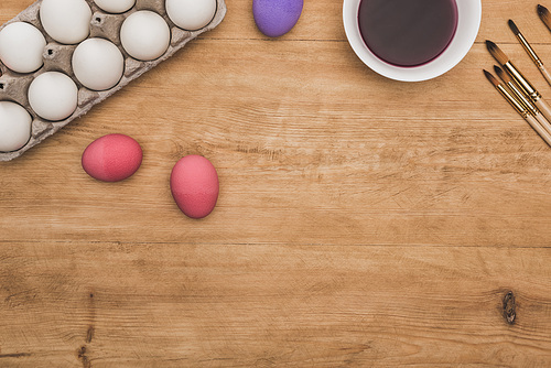 top view of watercolor purple paint in bowl near painted chicken eggs and paintbrushes on wooden table