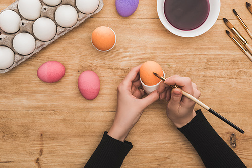 cropped view of woman coloring Easter eggs with paintbrush at wooden table