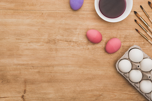 top view of watercolor purple paint in bowl near chicken eggs and paintbrushes on wooden table