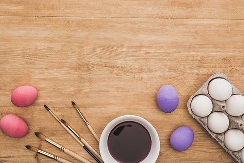 top view of watercolor purple paint in bowl near chicken eggs and paintbrushes on wooden table