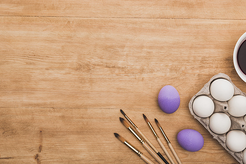 top view of watercolor purple paint in bowl near chicken eggs and paintbrushes on wooden table