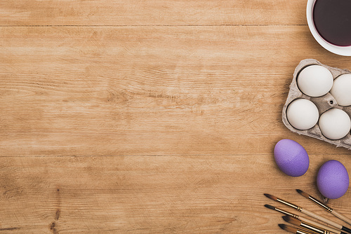 top view of watercolor purple paint in bowl near chicken eggs and paintbrushes on wooden table