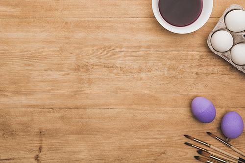 top view of watercolor purple paint in bowl near chicken eggs and paintbrushes on wooden table