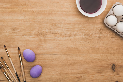 top view of watercolor purple paint in bowl near chicken eggs and paintbrushes on wooden table